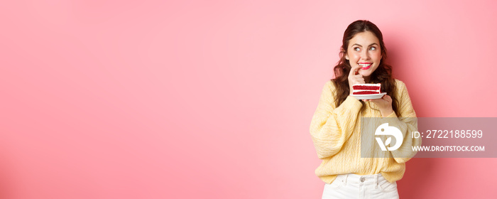 Cute glamour girl thinking and looking aside, wants to try delicious cake, holding plate with dessert and smiling, standing against pink background