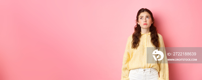 Image of upset and gloomy young woman pulling sad face, looking up with regret or jealousy, standing disappointed against pink background