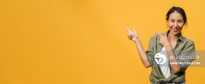 Young Asian lady smiling with cheerful expression, shows something amazing at blank space in casual cloth and looking at camera isolated over yellow background. Panoramic banner with copy space.