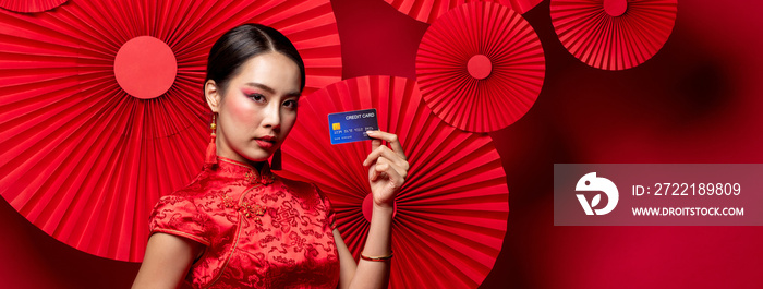 Portrait of beautiful Asian woman wearing traditional cheongsam qipao dress showing credit card in hand on red oriental banner background for Chinese new year shopping concepts