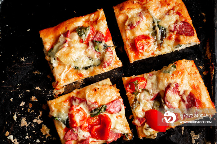 Sliced square pieces of pizza, pie, with tomatoes, sausage, cheese cooked in the oven on a black baking sheet.