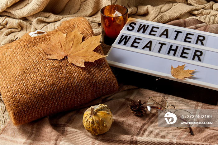 season and objects concept - wool clothes, autumn leaves, glasses and light box with sweater weather letters on warm blankets