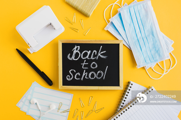 Blackboard with written text  back to school , protective medical face masks and school stationery on yellow background. Education during Covid 19 pandemic. Flat lay