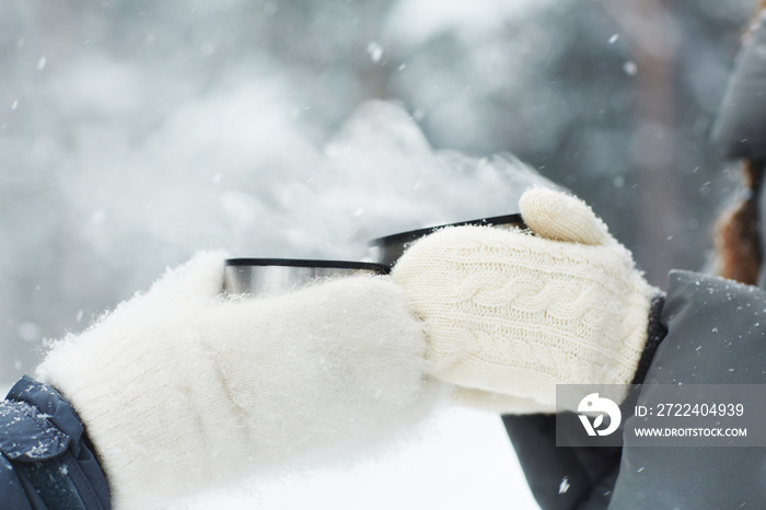 Two friends in white fluffy knitted mittens clinking with cups with hot drinks on snowy winter day outdoors