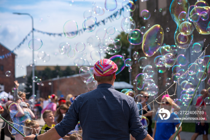 A freelance clown blowing hundreds of tiny, small and big bubbles at outdoor festival in city center. Concept of entertainment, birthdays. Kids having fun. Shower of bubbles flying in the happy crowd