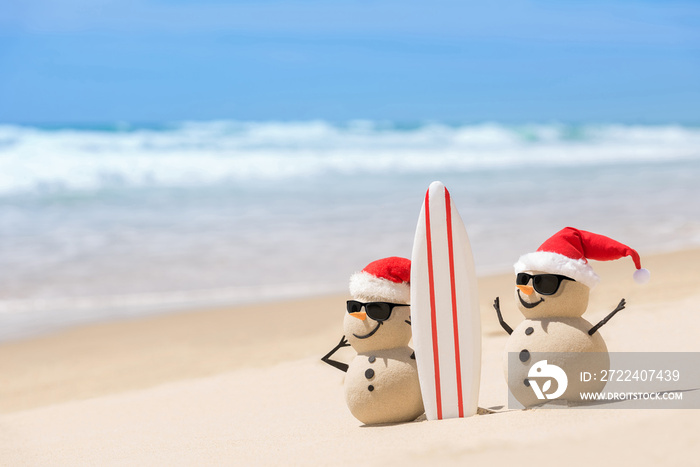 Two Sandy Christmas Snowmen are watching the waves, standing on beautiful beach with a surf board, wearing sunnies