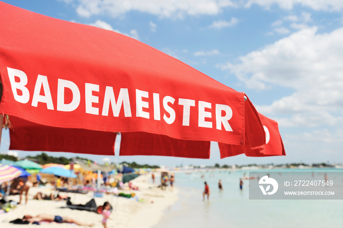 Red lifeguard umbrella on a beach against blue sky. Some bathers ont he background. The text on the umbrella Bademeister translates as Lifeguard, for german bathers. Risk and rescue concept.