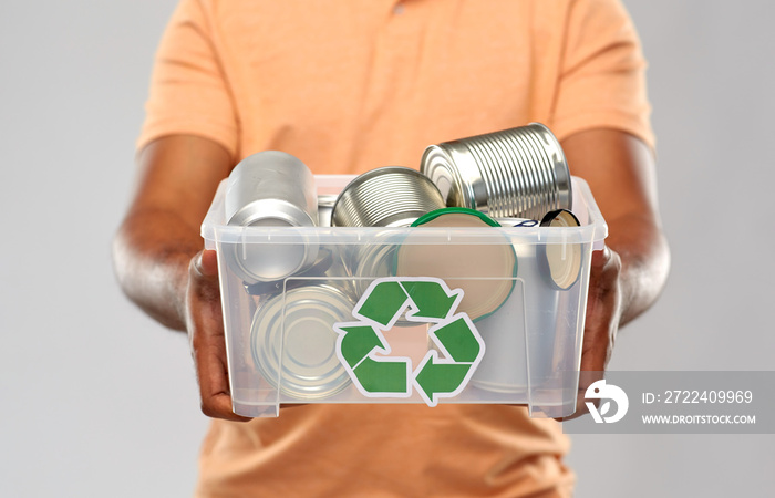 metal recycling, waste sorting and sustainability concept - close up of african american young man holding plastic box with tin cans over grey background