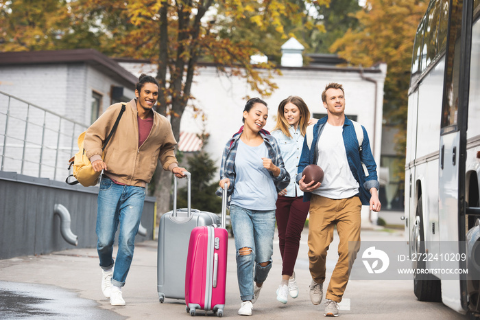 selective focus of smiling multiethnic tourists with wheeled bags and rugby ball running near travel bus at city street