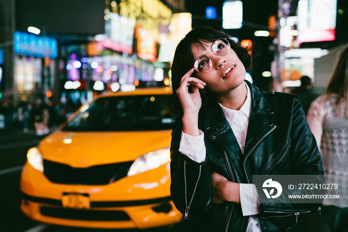 Charming brunette young woman in stylish spectacles looking up thinking at urban setting of night city,pensive hipster girl in leather jacket pondering about nightlife in metropolis standing at street