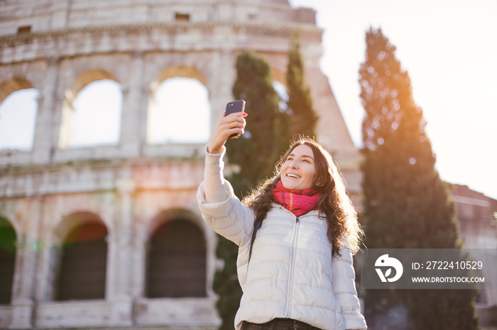 Young beautiful smiling woman posing against Roman colosseum and making selfie with smartphone - winter holidays and vacation in Europe concept