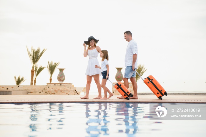 young family with suitcases walking to hotel building with beautiful swimming pool. traveling and relaxing time concept.