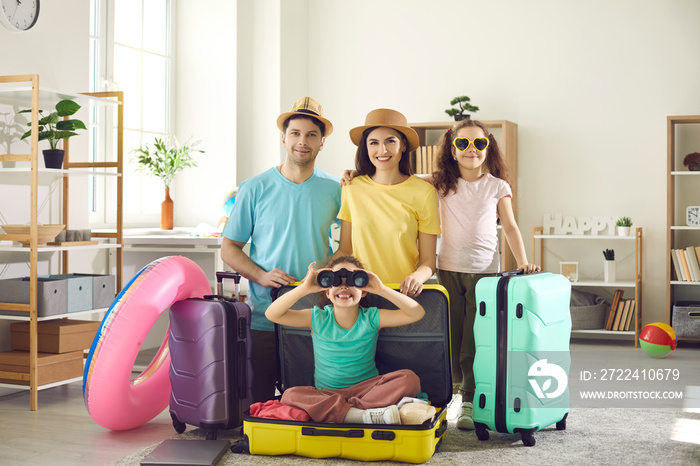 Ready to go on holiday. Portrait of happy family with travel cases packed for vacation trip. Mom, dad and kids smiling at camera, younger daughter sitting inside suitcase looking through binoculars