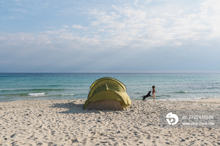 Camping tent on the beach and woman at distance running on the shore with her dog