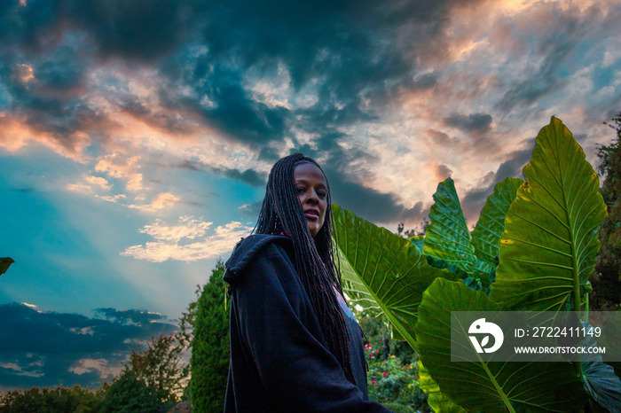 an African American woman with long sisterlocks wearing black and orange standing in the garden surrounded by lush green trees, plants and grass and colorful flowers with powerful clouds at sunset