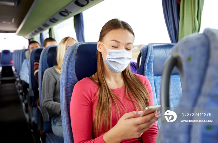 transport, tourism, road trip and people concept - young woman wearing face protective medical mask for protection from virus disease sitting in travel bus with smartphone