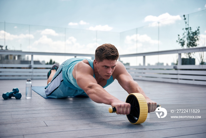 Concentrated young guy is exercising with sport equipment on roof of urban building. He is kneeling on mat and rolling forward outfit wheel for working on abdominal muscles. He is going to continue