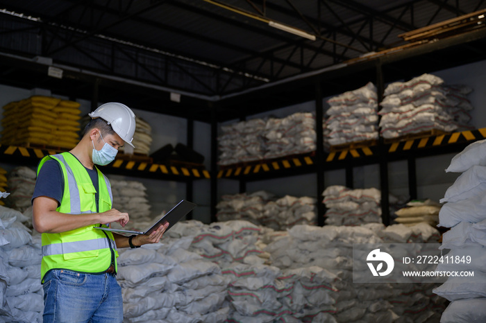 A male worker using a tablet computer working in an industrial chemical or parcel factory wears a white helmet in an industrial warehouse. The background is a warehouse and goods.