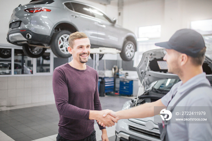 Happy men stand in garage and shake each others hands. They stand in front of opened body of car. There is another vehicle behind blonde man on platform.