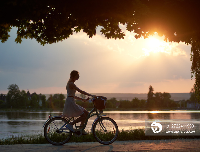 Happy woman riding a retro bike with a basket on the road near the lake on a sunset. Girl enjoying the magical view of the sun’s rays of the falling sun at the end of the day