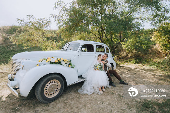 Loving newlyweds are sitting and hugging near an old retro car and summer nature. Wedding portrait of a stylish, smiling groom and pretty bride with curly hair. Photography and concept.