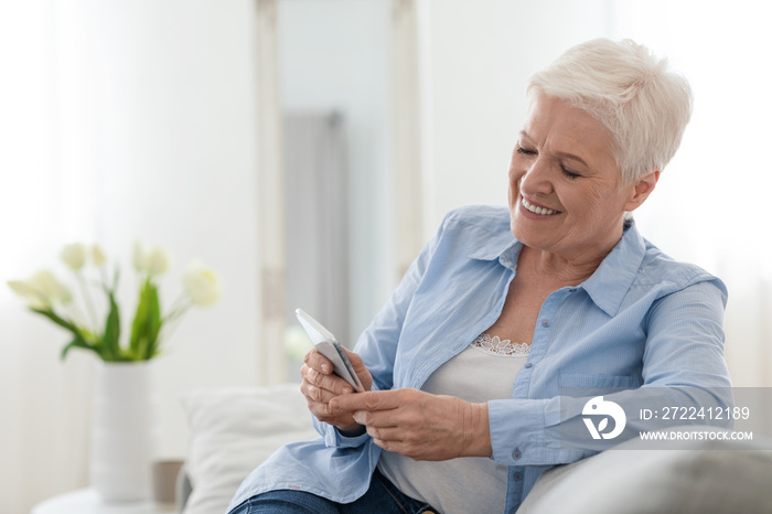 Happy elderly lady using mobile phone while relaxing on couch at home