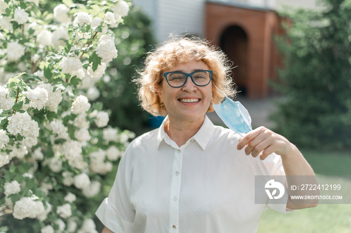 Happy old woman takes off the mask and sniffs a blossoming apple tree. A female pensioner enjoys fresh air on a warm summer day in the garden. Quarantine is over. The end of the coronavirus pandemic.