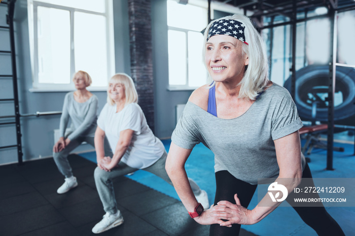 No time for worries. Selective focus on a smiling retired woman doing lunging exercises while standing in a row with a group of senior ladies training in a gym.