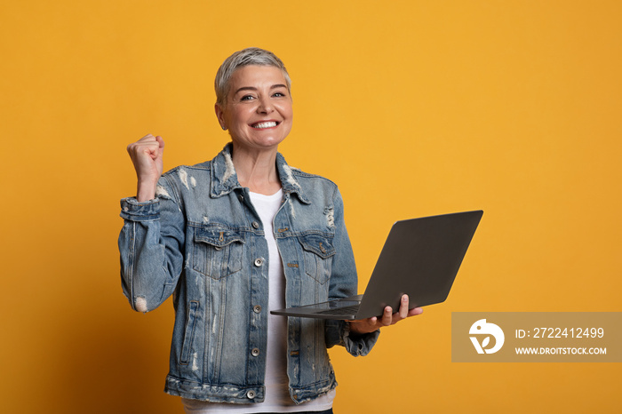 Joyful Middle-Aged Woman Holding Laptop, Celebrating Success Over Yellow Background