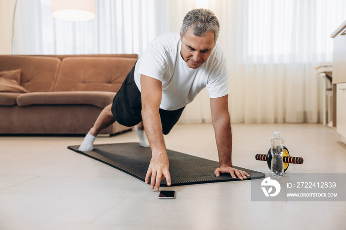 Elderly man reaches for phone to change music during excercise. Workout on yoga mat at home. Old man stands in plank position. Morning exercises with music. Active and happy old age during quarantine.