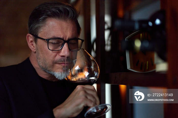 Middle aged man tasting, drinking wine in cellar. Wine expert rating a glass of red wine. Professional sommelier satisfied.