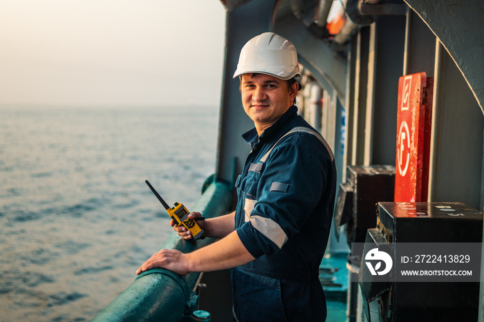 Marine Deck Officer or Chief mate on deck of offshore vessel or ship , wearing PPE personal protective equipment - helmet, coverall. He holds VHF walkie-talkie radio in hands.