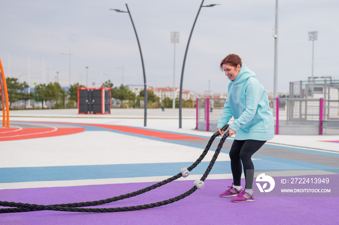 Caucasian woman in a mint sweatshirt is training with battle ropes at the sports ground.