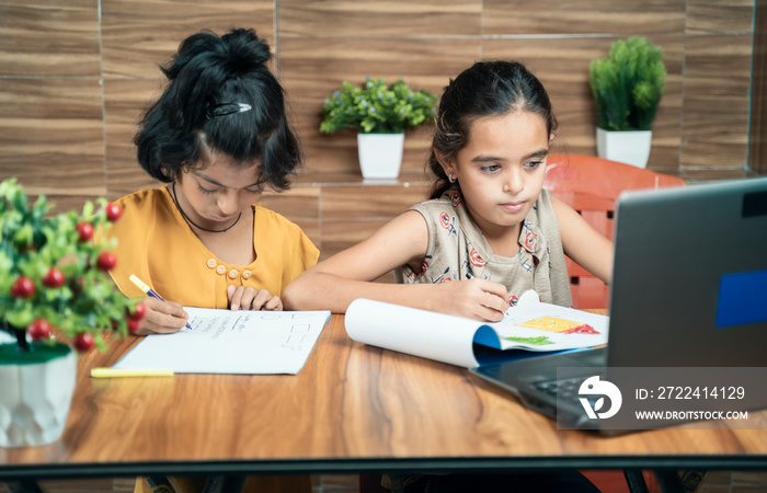 Two kids busy in writing by looking into the laptop during online or virtual class at home - concept of e-learning or distance learning or children lifestyle during coronavirus or covid-19 pandemic