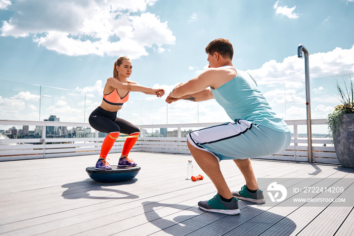 Strong man is doing sit-ups while girlfriend is squatting on fitness half ball and using resistance band on thighs. They are having joint work out on sunny terrace in city center under blue sky