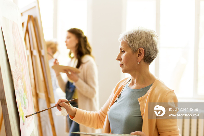 creativity, education and people concept - senior woman with brush and palette painting still life picture on easel at art school studio