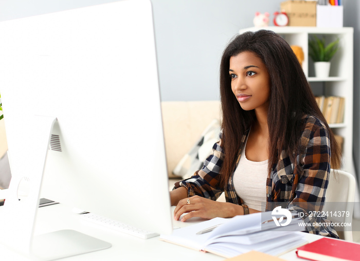 Black smiling woman sitting at workplace working with desktop pc studying or doing school homework concept