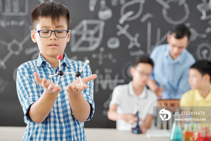 Curious schoolboy in glasses looking at plastic molecular model in his hands