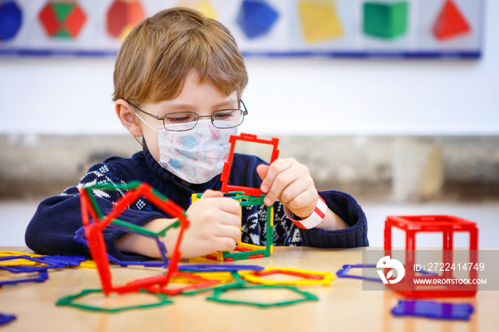 Little child with medical mask playing with lots of colorful plastic blocks kit in preschool nursery or elementary school. Cute child use protective equipment as fight against covid 19 corona virus.