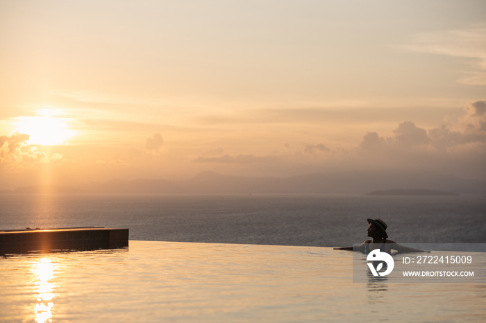 A woman with hat relaxing in infinity swimming pool and looking at a beautiful sunset and the sea view