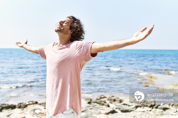 Young hispanic man breathing standing with arms open at the beach.