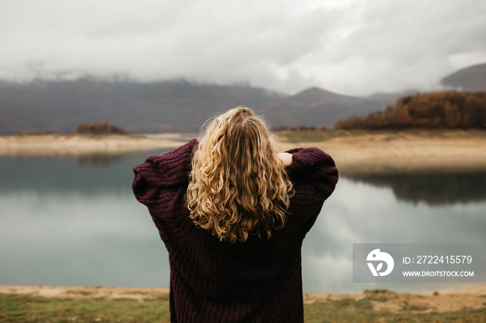 happy girl with curly blond hair dances on a lake alone, her hair is flying because of the wind flow, free as a bird. photo of girl with curly hair standing back to camera over lake background.