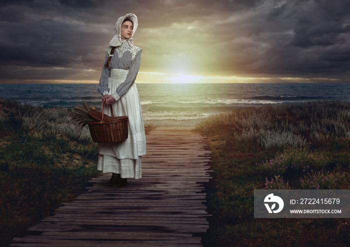 Victorian girl with a hood and a wicker basket containing dried flowers in a white dress and blue striped blouse on the coast at sunset.