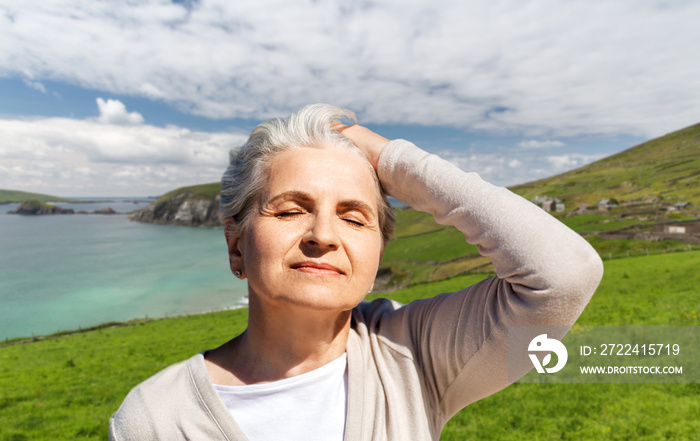 travel, tourism and vacation concept - portrait of happy senior woman enjoying sun over atlantic ocean coast in ireland background