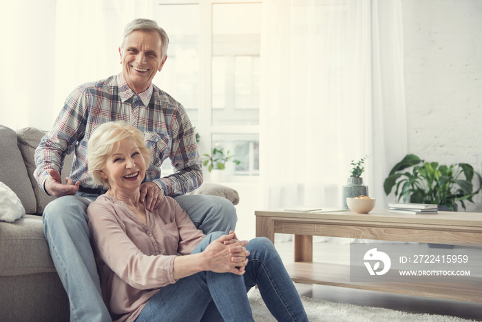 Portrait of content senior couple enjoying home comfort. Man is sitting on sofa and woman relaxing on the floor close to him. Copy space in right side