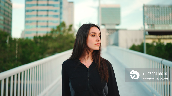 Portrait of cute girl with freckles and loose brown hair wearing black sports hoodie looks at the camera and walks on the bridge of the modern city background