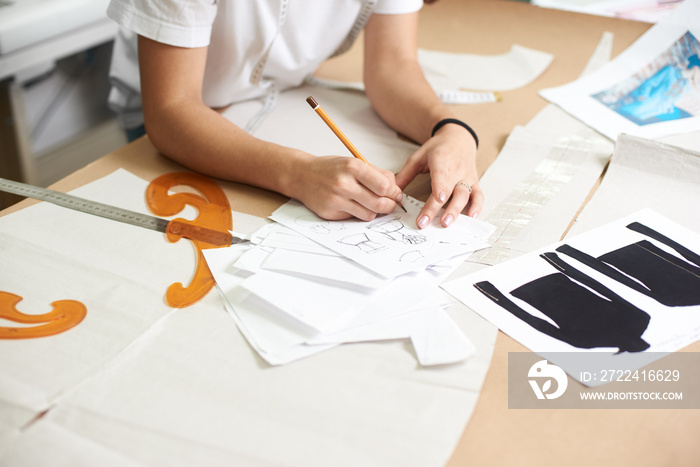 Female designer drawing pencil sketches of clothing sitting at big table with flat paper patterns, measuring ruler and curves on blurred background of tailor design studio interior.