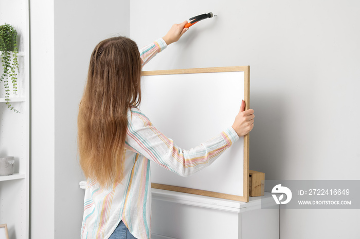 Young woman with blank frame nailing on light wall at home