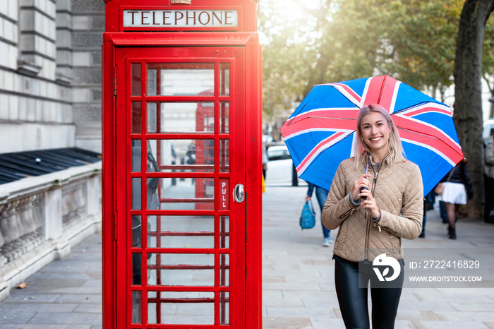 Reise Konzept für London: glückliche, blonde Frau mit Regenschirm aus britischer Fahne vor einer klassisch, roten Telefonzelle, Großbritannien