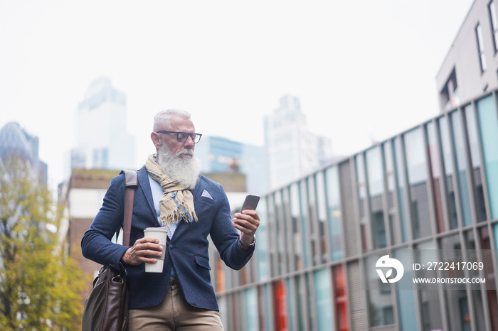 Senior hipster man using mobile phone and drinking coffee with city in background - Focus on face